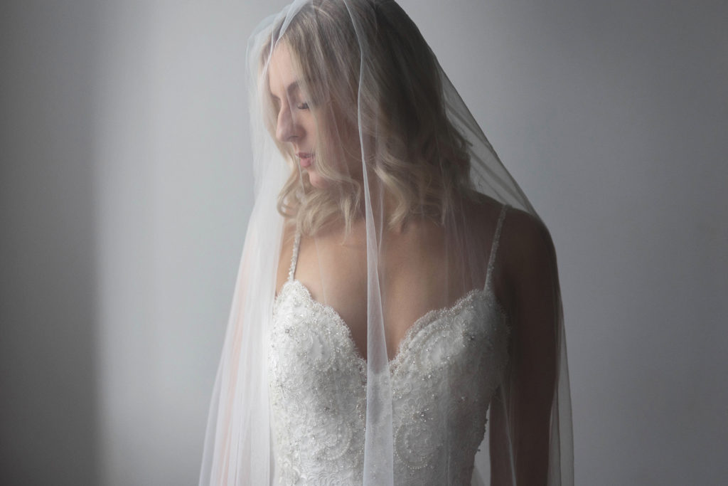 Close up of bride standing in a white room, veil over her right shoulder, strap of her dress fallen down on her left shoulder. She is looking down to the left.